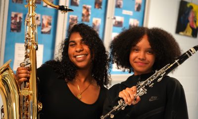 Two young women with black curly hair hold their instruments up and smile. One has a saxophone, the other a clarinet.