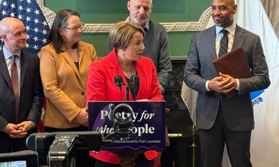 photo of Governor Healey speaking at a podium to press. Behind her stand four esteemed guests looking on. In the foreground a video camera slightly obscures a sign on the podium that reads Poetry for the People.