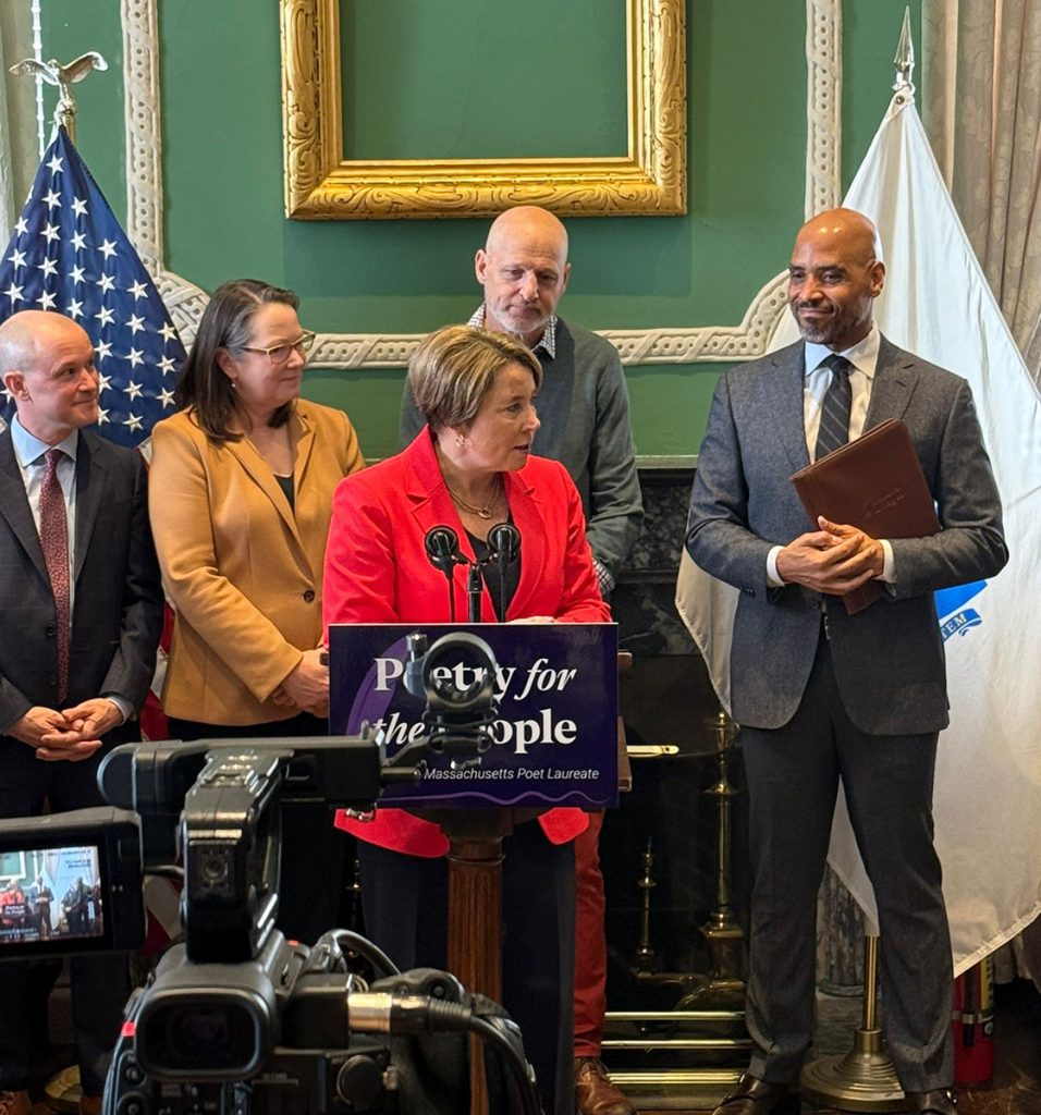 photo of Governor Healey speaking at a podium to press. Behind her stand four esteemed guests looking on. In the foreground a video camera slightly obscures a sign on the podium that reads Poetry for the People.