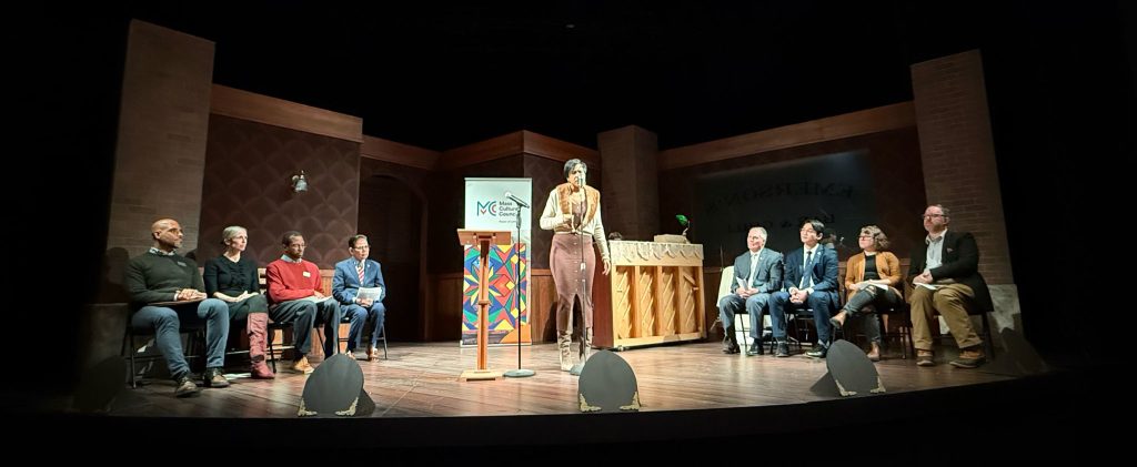 photo of a stage with four speakers seated on both sides of a woman who delivering a monologue.