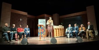 photo of a stage with four speakers seated on both sides of a woman who delivering a monologue.