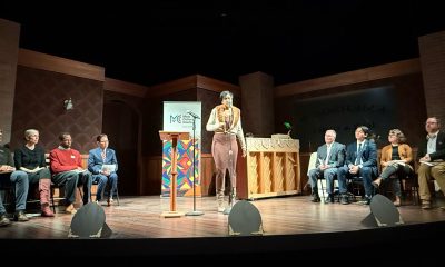 photo of a stage with four speakers seated on both sides of a woman who delivering a monologue.