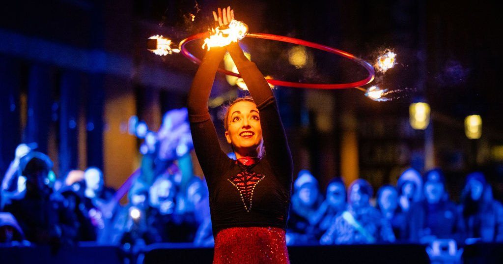 photo of a fire artist twirling a hoop over her head with some flames on it. Behind her is a a crowd of onlookers watching in the otherwise dark night.