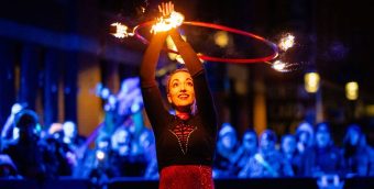 photo of a fire artist twirling a hoop over her head with some flames on it. Behind her is a a crowd of onlookers watching in the otherwise dark night.