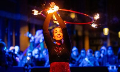 photo of a fire artist twirling a hoop over her head with some flames on it. Behind her is a a crowd of onlookers watching in the otherwise dark night.