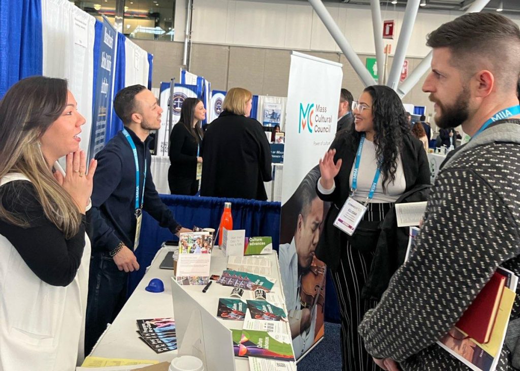 photo of two Mass Cultural staff standing on one side of a long table covered in pamphlets speaking across it to two conference attendees.