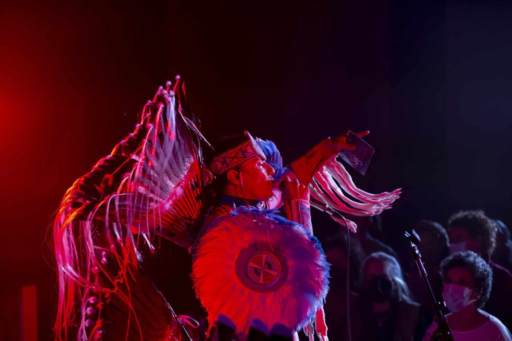 photo of a rapper in blue and white Native American garb seen from the side. He's holding a mic in one hand and point the other out into the audience.