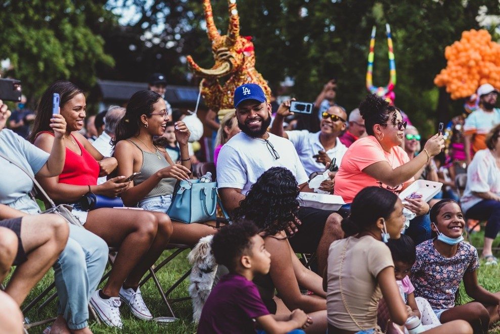 photo of a crowd of people sitting outside on a warm day. Some are in folding chairs and some are sitting on the grass.