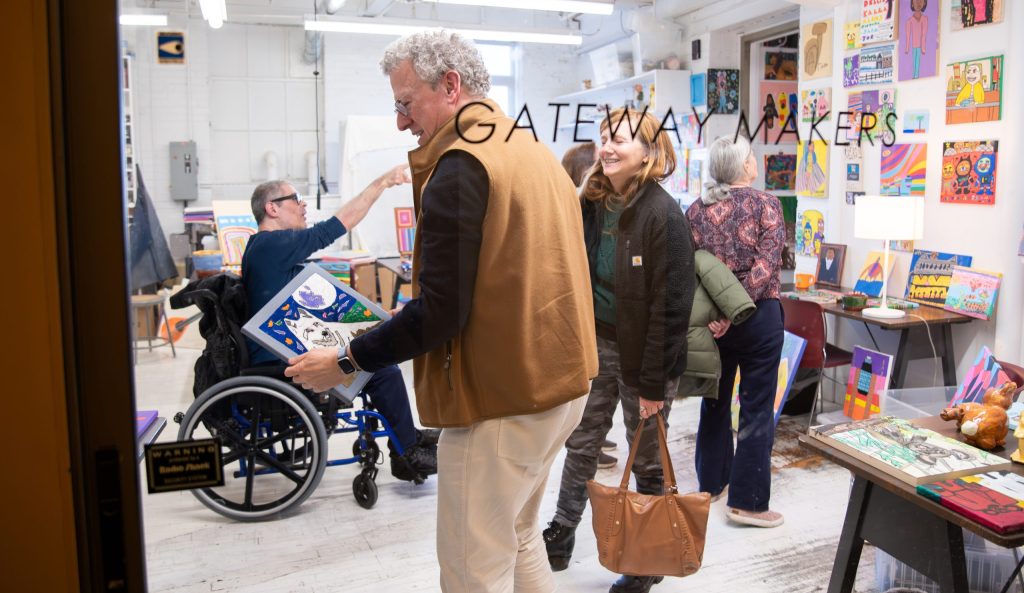 photo looking through a glass interior wall with the words "Gateway Makers". On the other side numerous people are holding art works and looking at pieces on display for purchase.