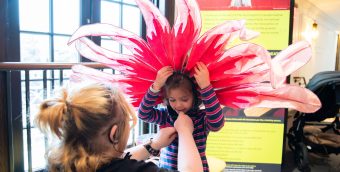 photo of a small child getting outfitted with a large pink and red feather head dress like you might see as part of Carnival.
