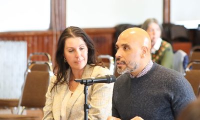 photo of a woman sitting next to a man who is speaking into a microphone.