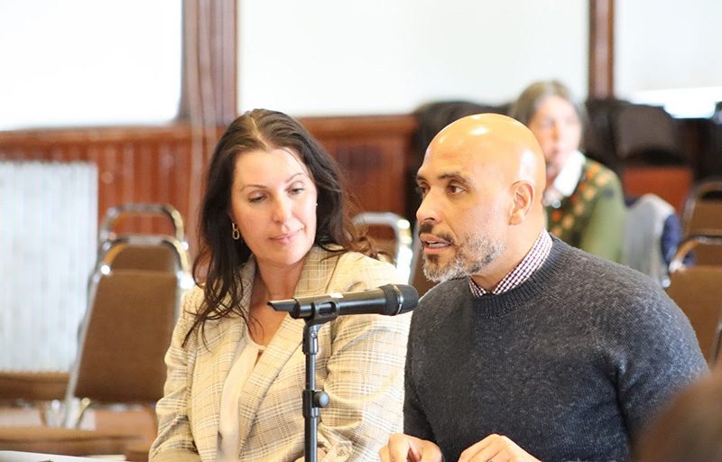 photo of a woman sitting next to a man who is speaking into a microphone. 