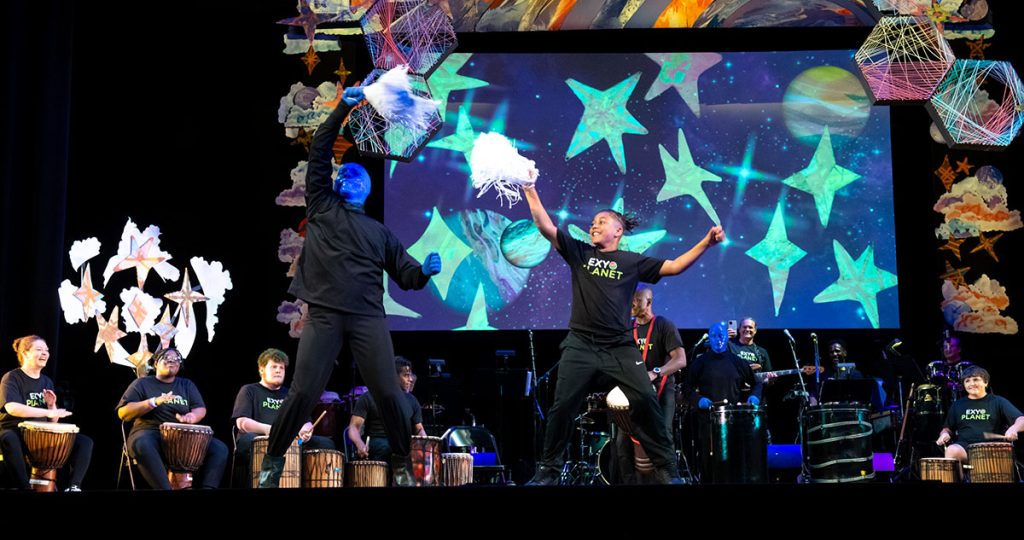 theater performance still of a stage with a performer from Blue Man Group waving a pom pom next to a young person also waving a pom pom. Behind them is a row of musicians, some youth and some adults, playing.