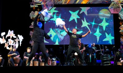 theater performance still of a stage with a performer from Blue Man Group waving a pom pom next to a young person also waving a pom pom. Behind them is a row of musicians, some youth and some adults, playing.