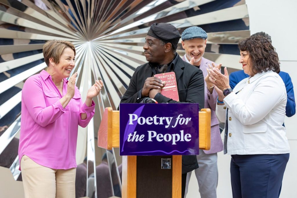 photo of a man at a podium. on either side of him are two woman clapping for him and smiling.
