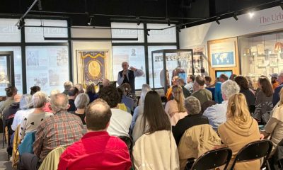 photo of a man in the background speaking to an audience seated in a history museum's gallery, their backs to the camera, in the foreground.