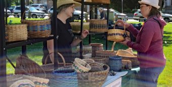 photo of a booth selling baskets at an outdoor festival. There's a woman selling the baskets speaking to a woman standing at the booth.
