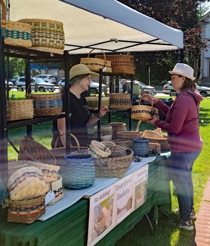 photo of a booth selling baskets at an outdoor festival. There's a woman selling the baskets speaking to a woman standing at the booth.