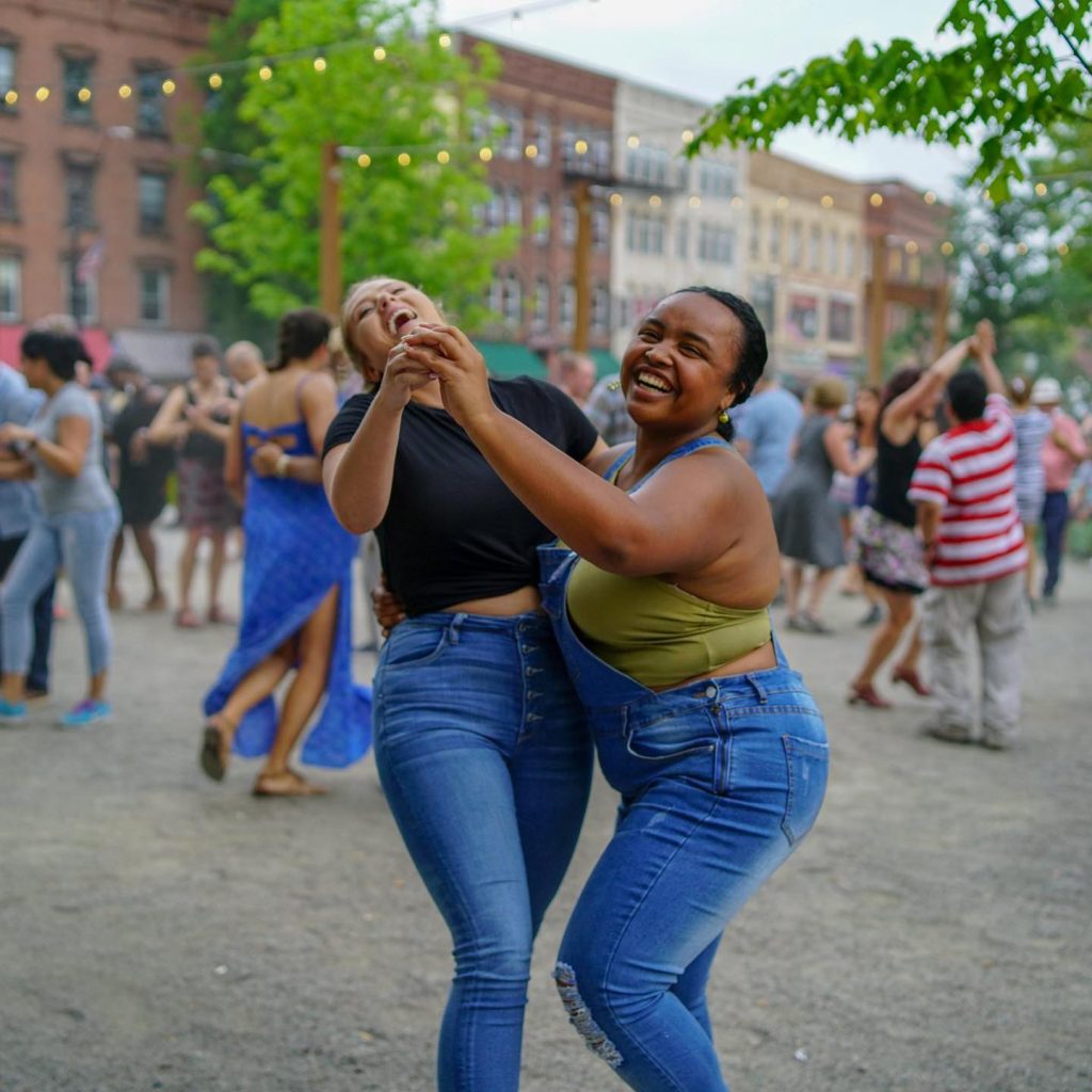 two women wearing warm-weather tops and blue jeans dancing Salsa together and laughing at an outdoor community dance
