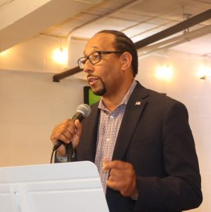photo of a man standing, speaking into a mic.