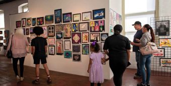 photo of an art gallery wall covered in small, tightly hung together pieces. A couple of visitors are looking at the art.