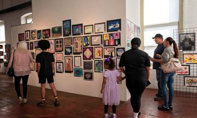 photo of an art gallery wall covered in small, tightly hung together pieces. A couple of visitors are looking at the art.
