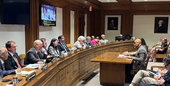 photo of a hearing room. a panel of legislators sits in a row listening to a seated man speaking into a microphone.