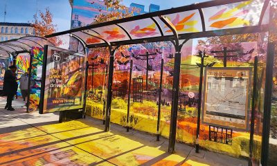 photo of a bus shelter with it's plexiglass top and sides painted in bright, warm yellows and oranges. the sun is pouring through it, creating a reflection of colors on the sidewalk in front of it.