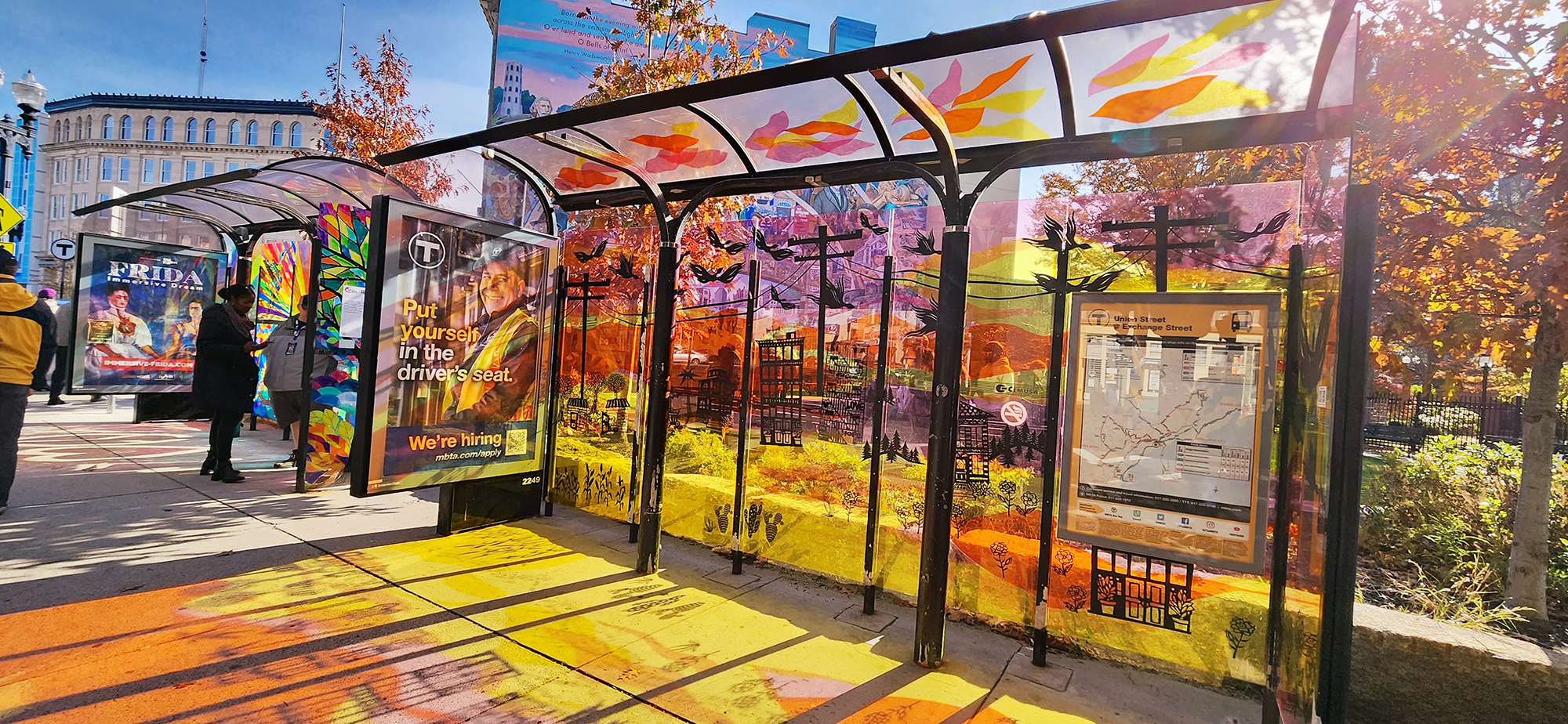 photo of a bus shelter with it's plexiglass top and sides painted in bright, warm yellows and oranges. the sun is pouring through it, creating a reflection of colors on the sidewalk in front of it.