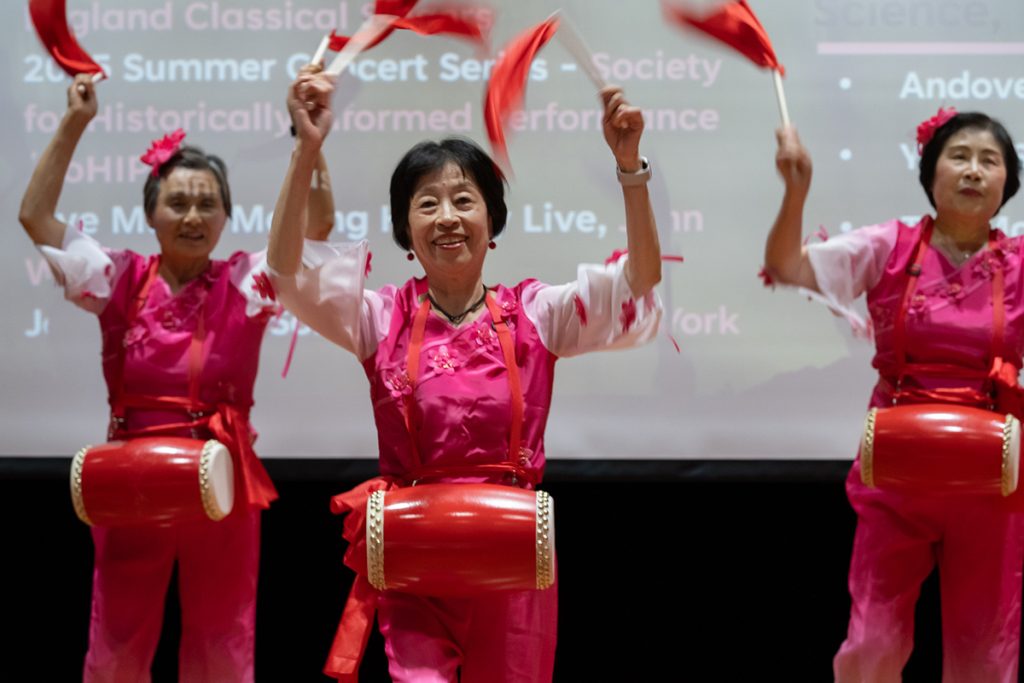 three older woman wearing pink and red have drums around their waists and wave red pendant flags above their heads.