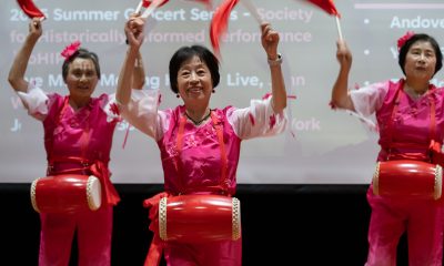 three older woman wearing pink and red have drums around their waists and wave red pendant flags above their heads.