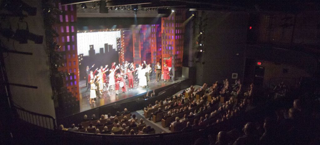 photo of the inside of a theater from the back left corner. The stage is illuminated with a production and the seats are filled.