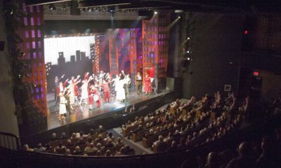 photo of the inside of a theater from the back left corner. The stage is illuminated with a production and the seats are filled.
