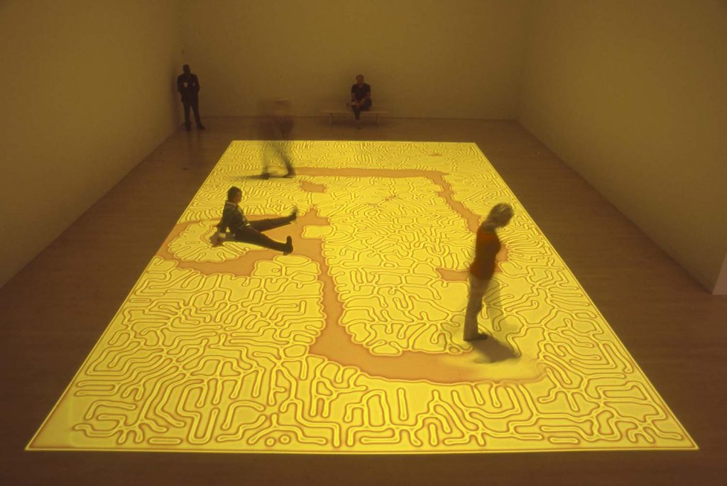 photo from a video installation with a bright yellow and orange projection on a gallery floor interrupted by visitors' walking through it, the yellow seeping back from where it had been interrupted.
