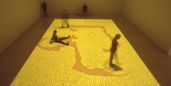 photo from a video installation with a bright yellow and orange projection on a gallery floor interrupted by visitors' walking through it, the yellow seeping back from where it had been interrupted.