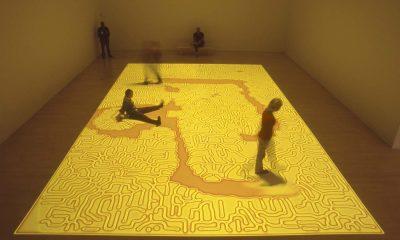 photo from a video installation with a bright yellow and orange projection on a gallery floor interrupted by visitors' walking through it, the yellow seeping back from where it had been interrupted.