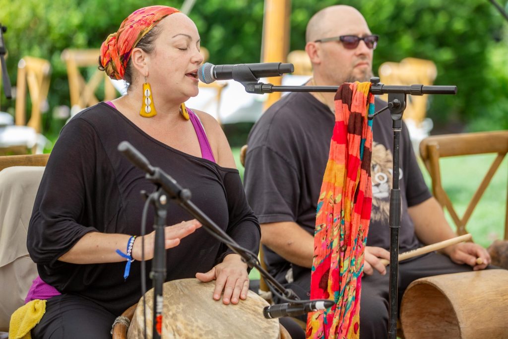 photo of a woman playing a barriles and singing into a microphone with a man standing next to her holding cuá.