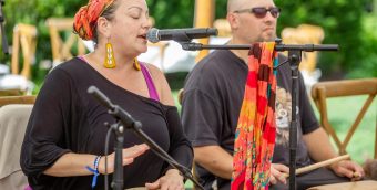 photo of a woman playing a barriles and singing into a microphone with a man standing next to her holding cuá.