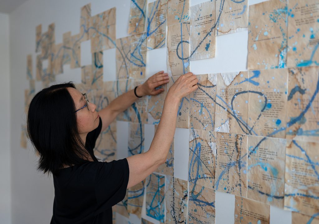 photo of an artist adjusting an installation in-progress in her studio. Dozens of pieces of yellowed typewritten papers are hung in an abstract design with blue swirls of paint overlaid.