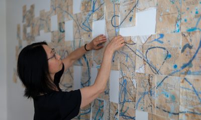 photo of an artist adjusting an installation in-progress in her studio. Dozens of pieces of yellowed typewritten papers are hung in an abstract design with blue swirls of paint overlaid.