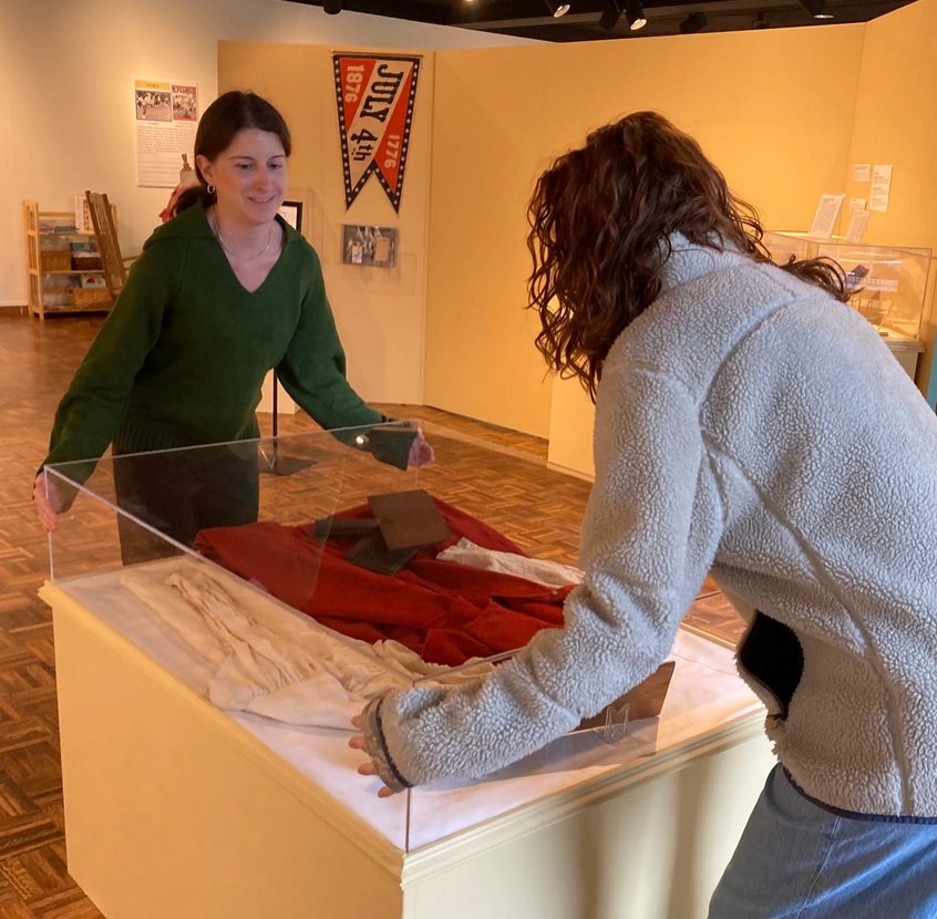 photo of two women in a gallery space adjusting a protective display case over an American Revolution era shirt and jacket. 