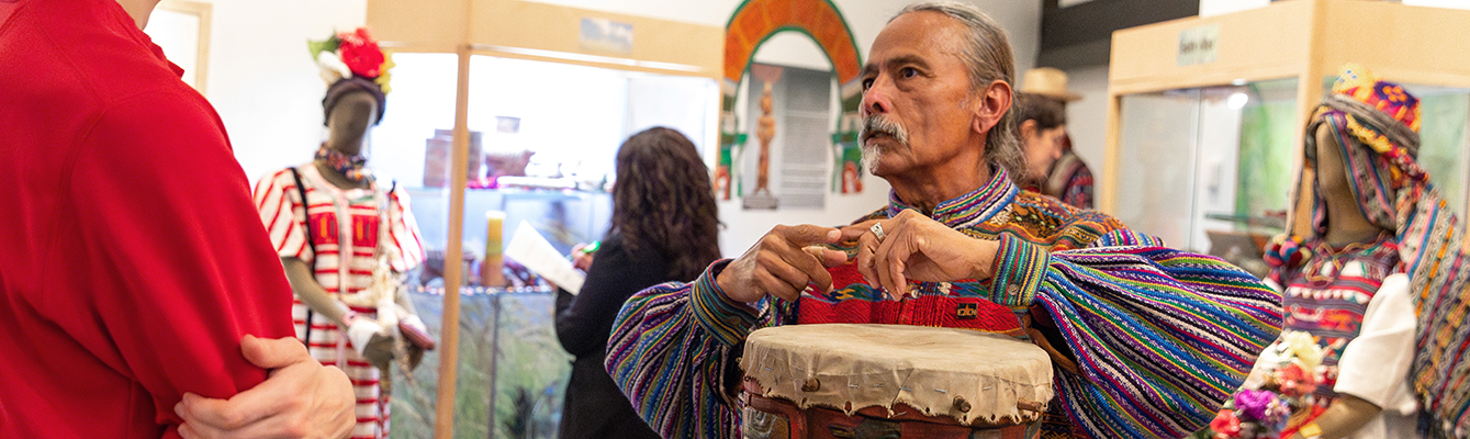photo of an Indigenous man wearing a traditional Mayan top, his long grey hair is pulled back. His arms are raised over an intricately carved wooden drum.