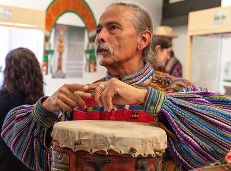 photo of an Indigenous man wearing a traditional Mayan top, his long grey hair is pulled back. His arms are raised over an intricately carved wooden drum.