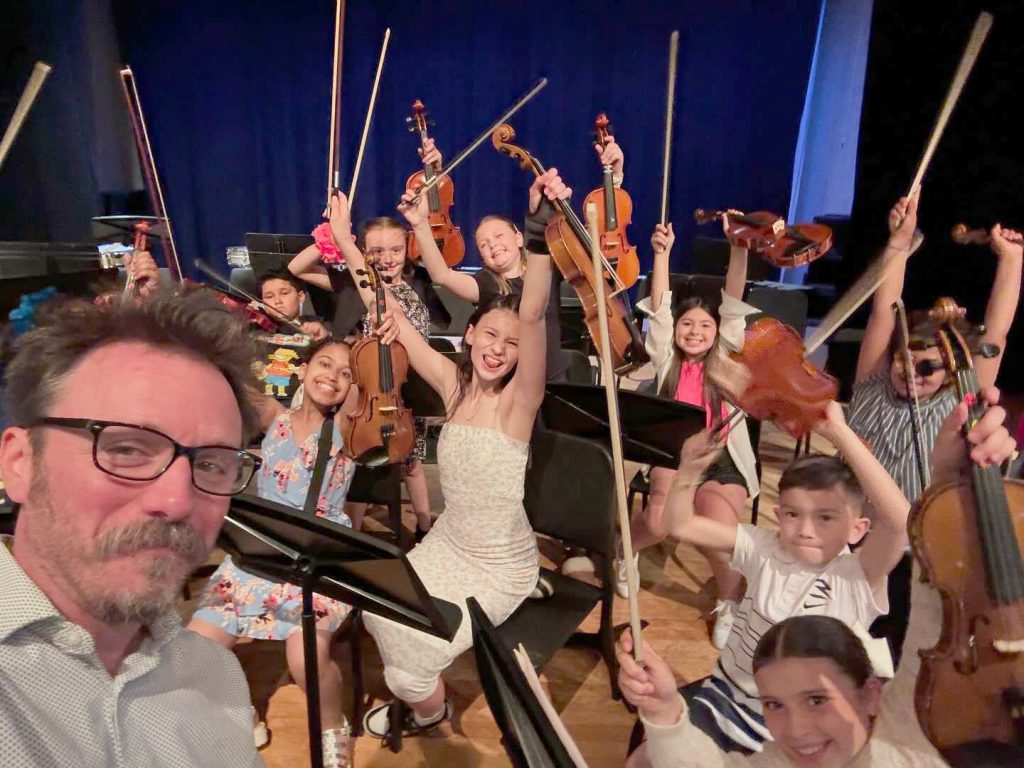 music teacher taking a selfie with a bunch of enthusiastic student musicians who are smiling have their arms and instruments raised in the air in celebration.
