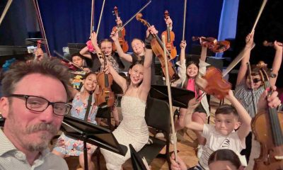 music teacher taking a selfie with a bunch of enthusiastic student musicians who are smiling have their arms and instruments raised in the air in celebration.