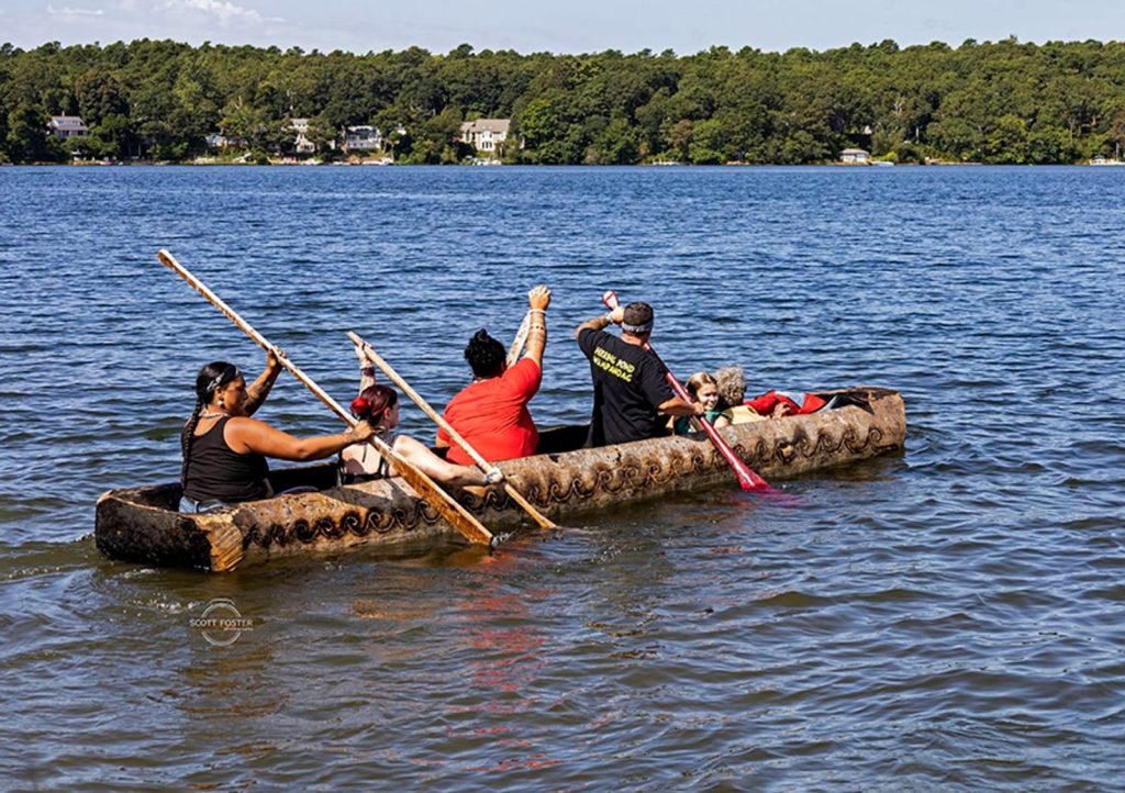 photo of a half dozen adults and children paddling in a canoe made by hand in the tradition of the Herring Pond Wampanoag Tribe.
