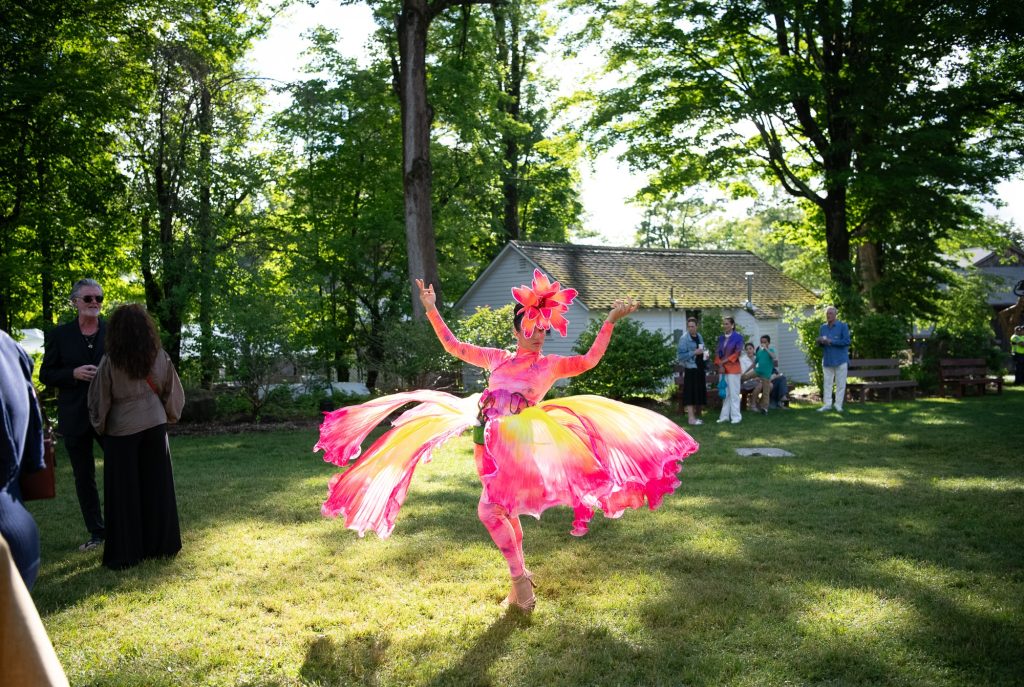 photo of a dancer costumed in vibrant pinks and yellows posed with their arms uplifted in a grassy space with lots of greenery and trees and onlookers in the background.
