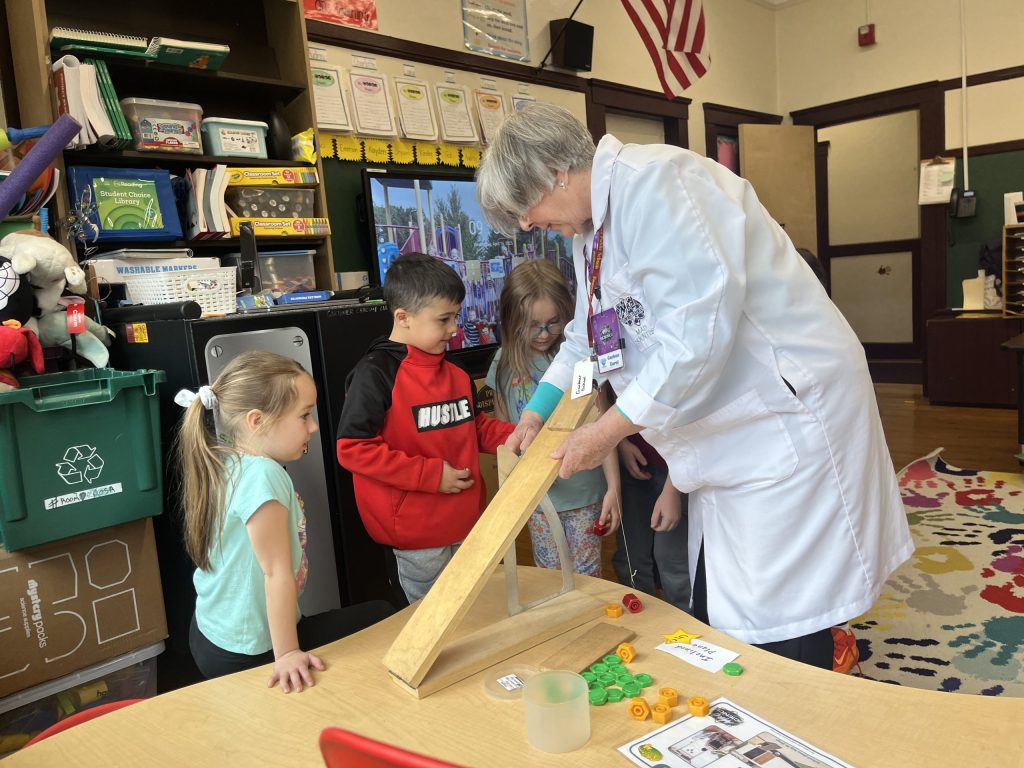 photo of an elementary school classroom where an older scientist is explaining something using a piece of wood held at an angle. 3 school children watching her closely.