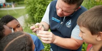 photo of a science educator holding a caterpillar while out in nature, a circle of curious students surrounds him.