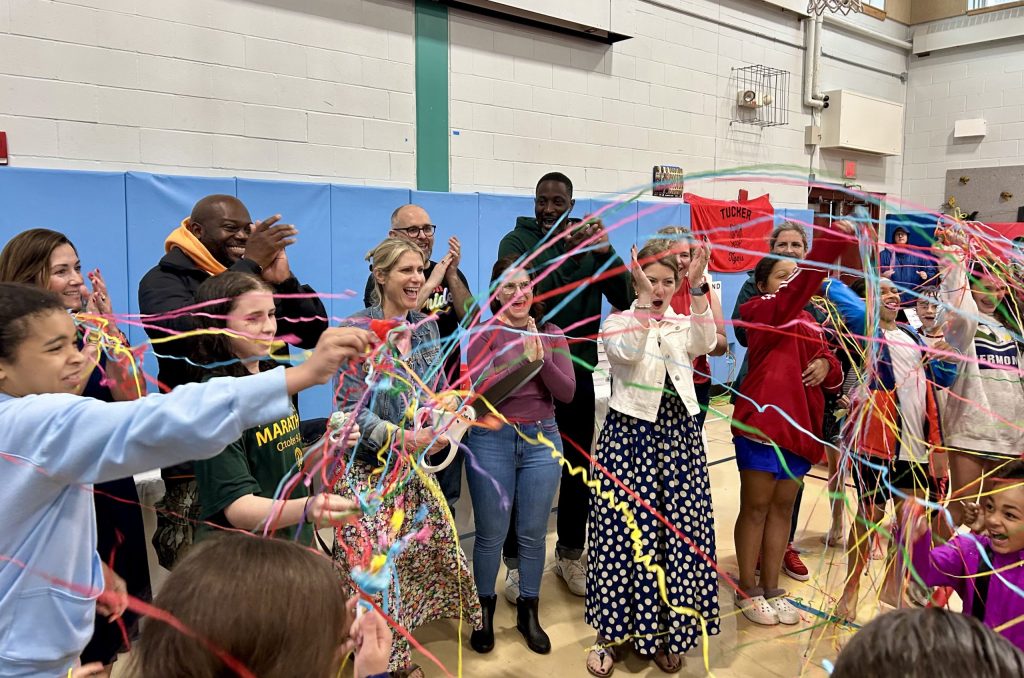 A teaching artist stands with a group of teachers and student in a school gym. Many of them are throw streamers and cheering.
